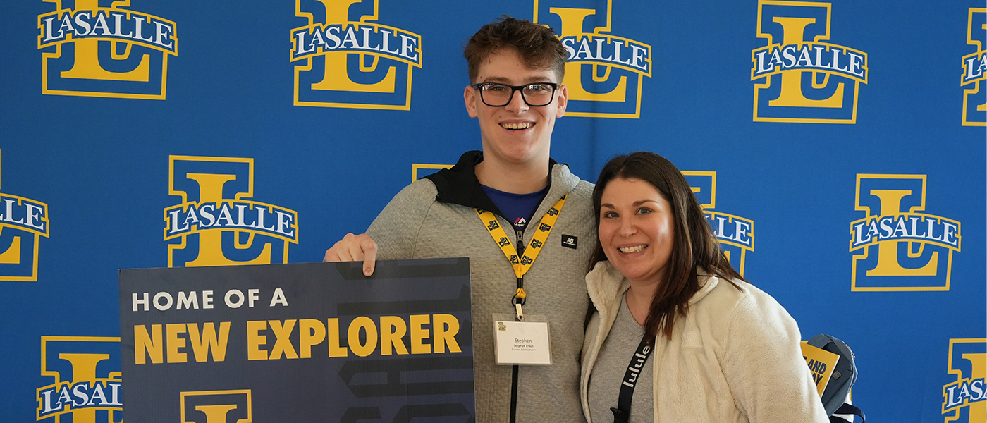Image of a male student holding a "Home of a New Explorer" sign with his mom.
